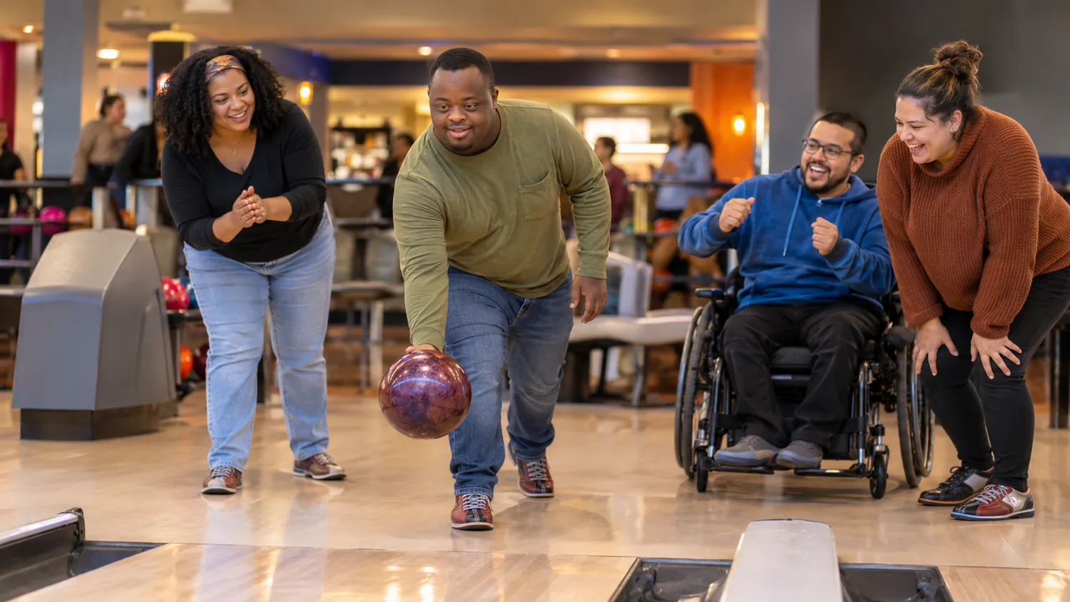 A man in a green shirt rolls a bowling ball down a lane while a woman in a black top, a man in a wheelchair, and a woman in a rust-colored sweater cheer him on.